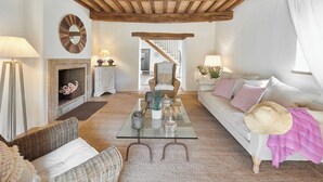 Een gezellige woonkamer met een open haard, rieten meubels, een glazen salontafel en een witte bank met opvallende kussens.