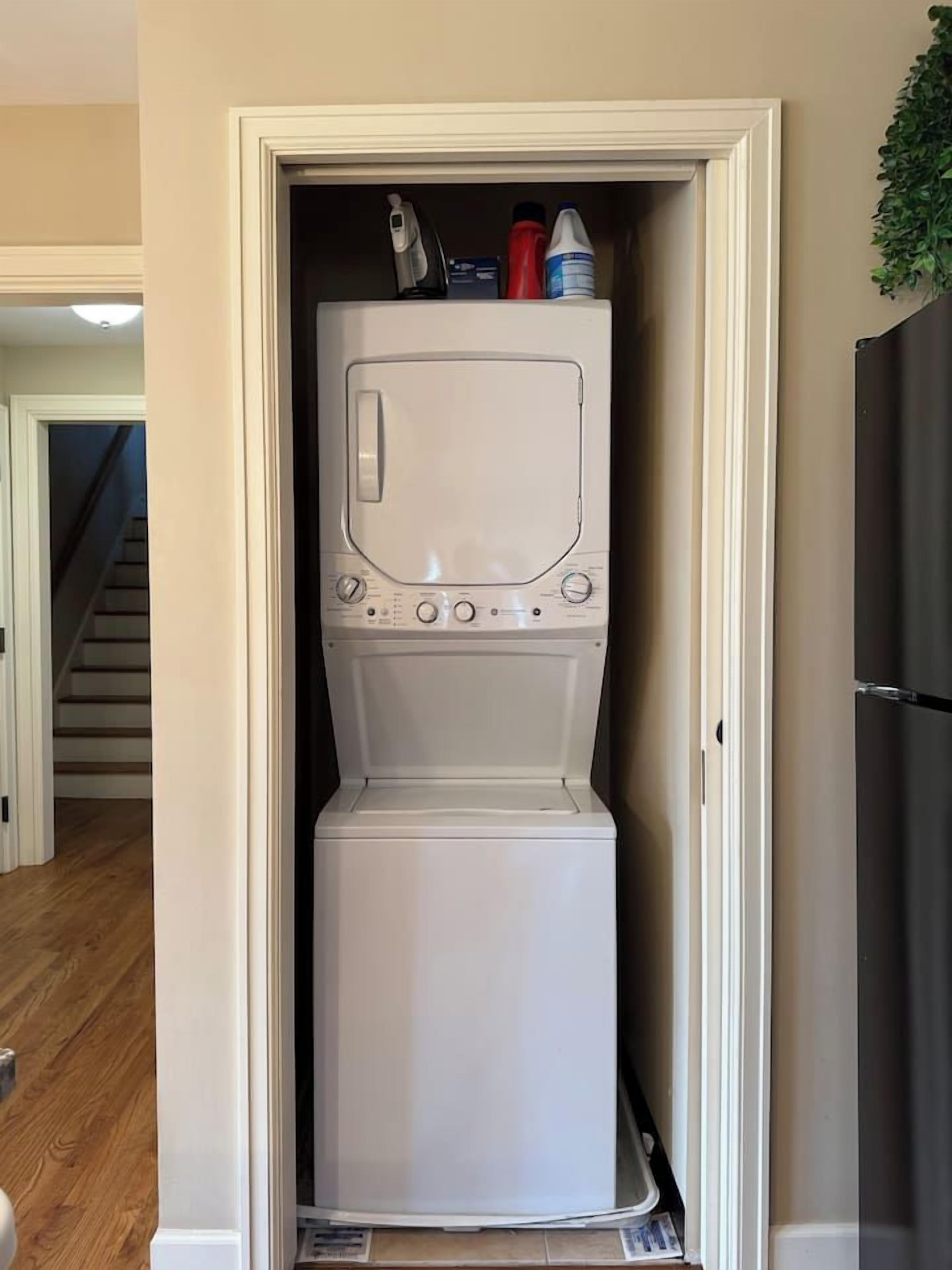 A laundry room with a front-loading washer and dryer, a small shelf with cleaning supplies, and a staircase in the background.