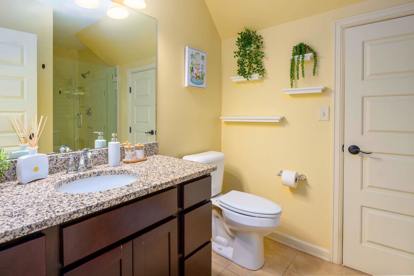 Une salle de bain avec un lavabo sur un plan de travail en granit, des toilettes et une douche. On y trouve des plantes sur des étagères murales et un tableau encadré au mur.