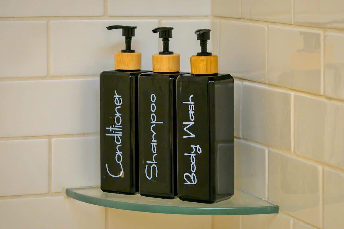 Three black bottles with yellow pumps labeled 'Conditioner,' 'Shampoo,' and 'Body Wash' on a glass shelf against a tiled wall.
