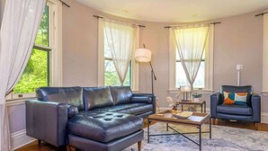 Un salón con un sofá de cuero, una mesa de centro y una silla. Hay dos ventanas con cortinas, una lámpara de pie y una mesita con una lámpara y objetos decorativos.