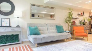 A living room with a grey sofa, teal cushions, a small orange chair, a white side table, a mirror, and a green plant.