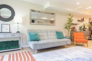 Una sala de estar con un sofá gris, cojines azul verdosos, un sillón naranja pequeño, una mesita blanca, un espejo y una planta verde.