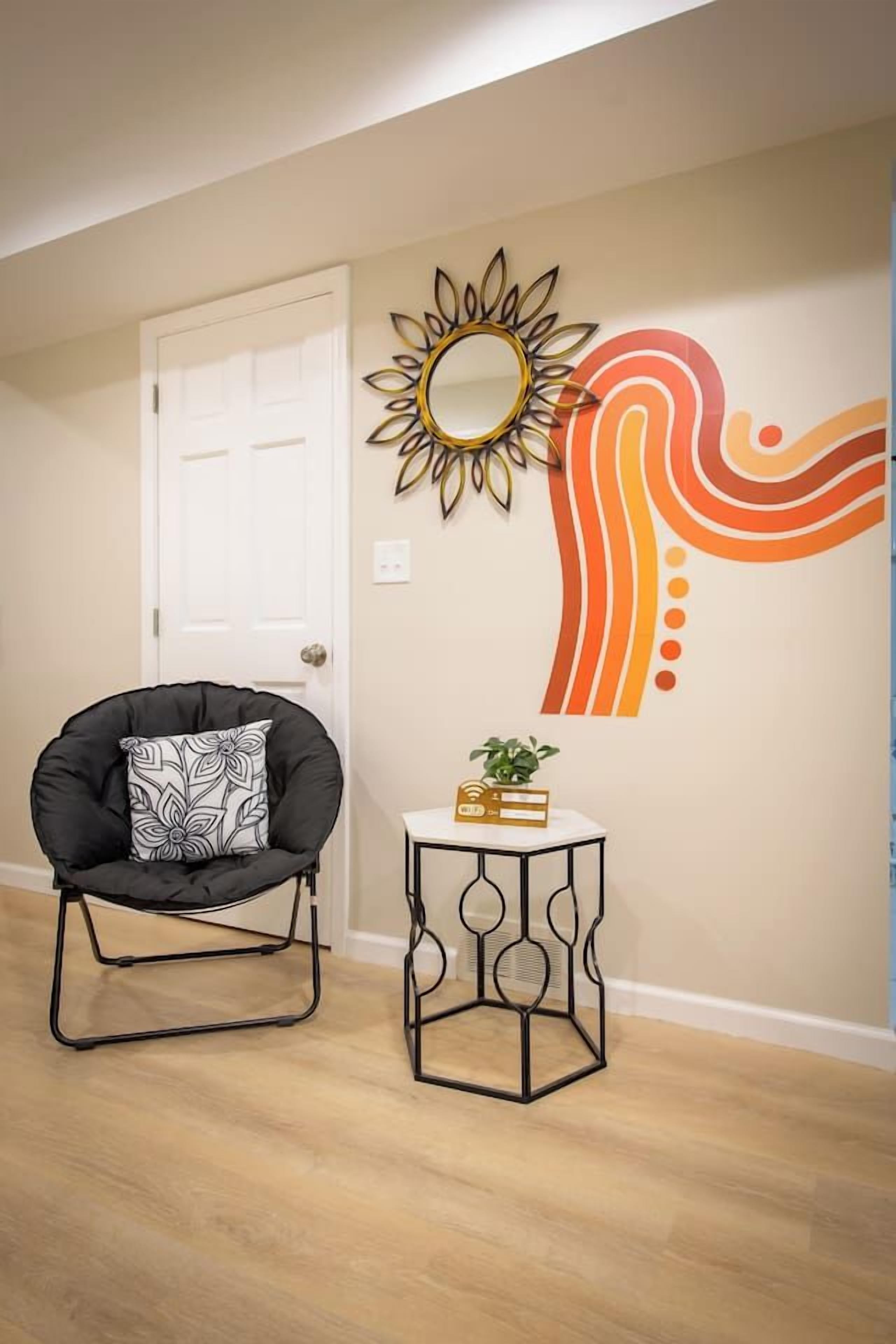 A room with a door, a black chair with a patterned pillow, a small round table with a plant, and a wall with a sunflower mirror and abstract art.