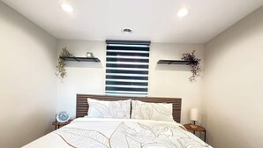 A bedroom with a large bed, a headboard with horizontal slats, bedside tables with lamps, and wall-mounted shelves with plants.