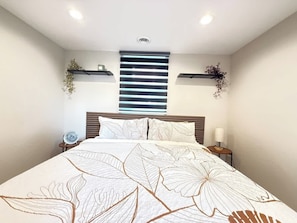 Un dormitorio con una cama grande, un cabecero con listones horizontales, mesitas de noche con lámparas y estantes en la pared con plantas.