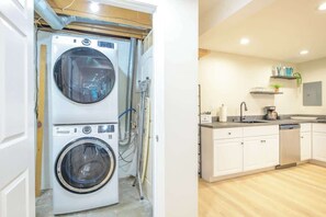 Un lavadero con lavadoras y secadoras apiladas, una cocina con armarios blancos y una encimera con diversos objetos.