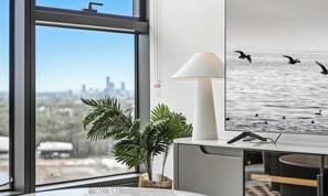 Ein modernes Büro mit großem Fenster, Schreibtisch, Lampe und einem an der Wand befestigten Bildschirm, der Vögel über Wasser zeigt.