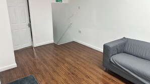 A room with a grey sofa, a wooden floor, a white door, and a green fire exit sign. - Two bed flat in Manchester—Walk to Uni & Hospitals (Manchester)