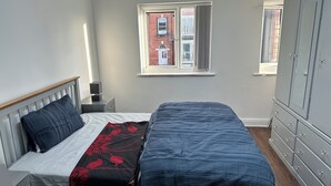 A bedroom with a bed, a nightstand, a dresser, and a window with a view of buildings. - Two bed flat in Manchester—Walk to Uni & Hospitals (Manchester)