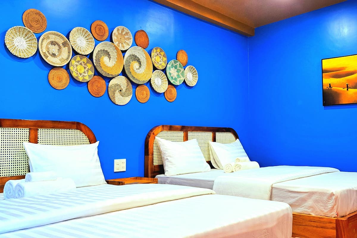 A hotel room with two beds, a blue wall, and a wall art of a desert sunset.