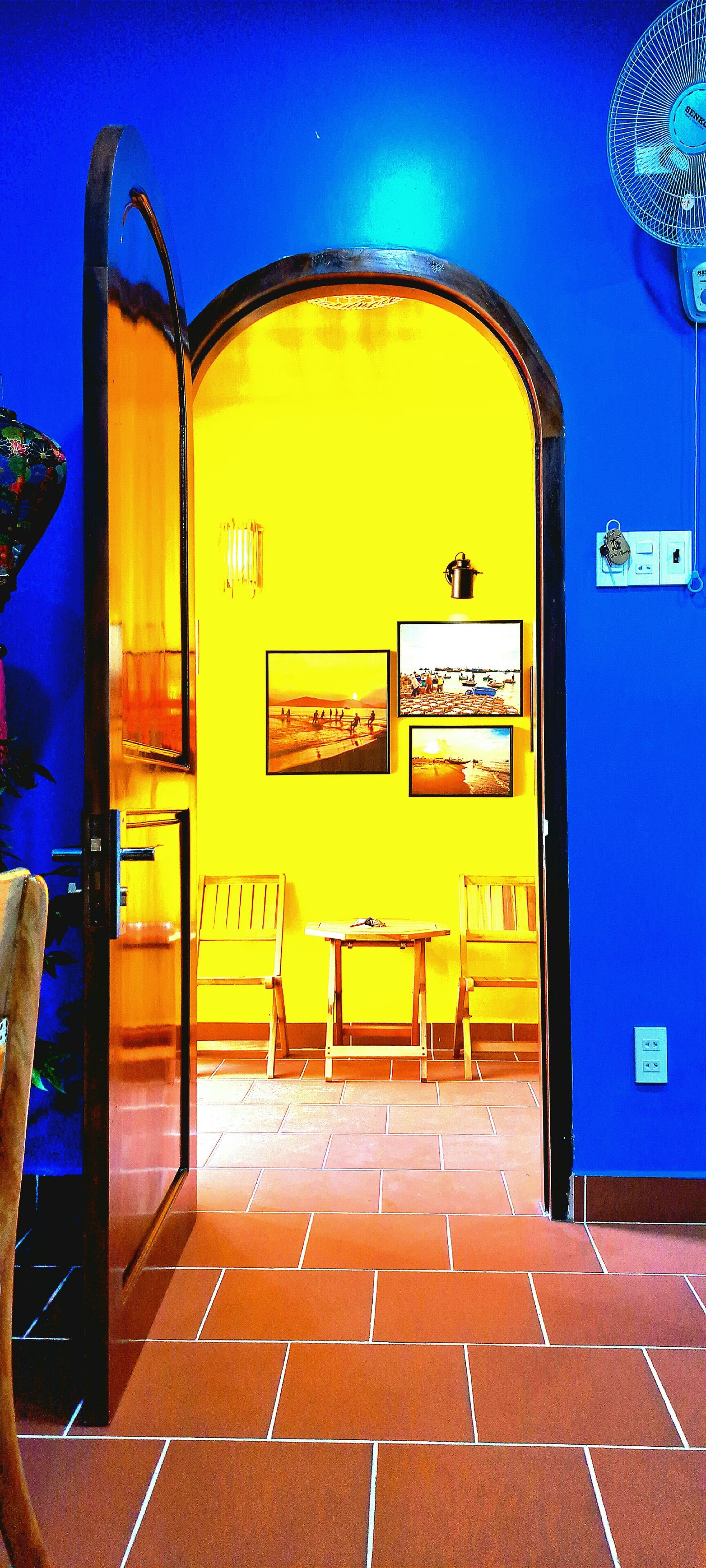 A room with a bright yellow wall, a wooden table, two chairs, and a fan.