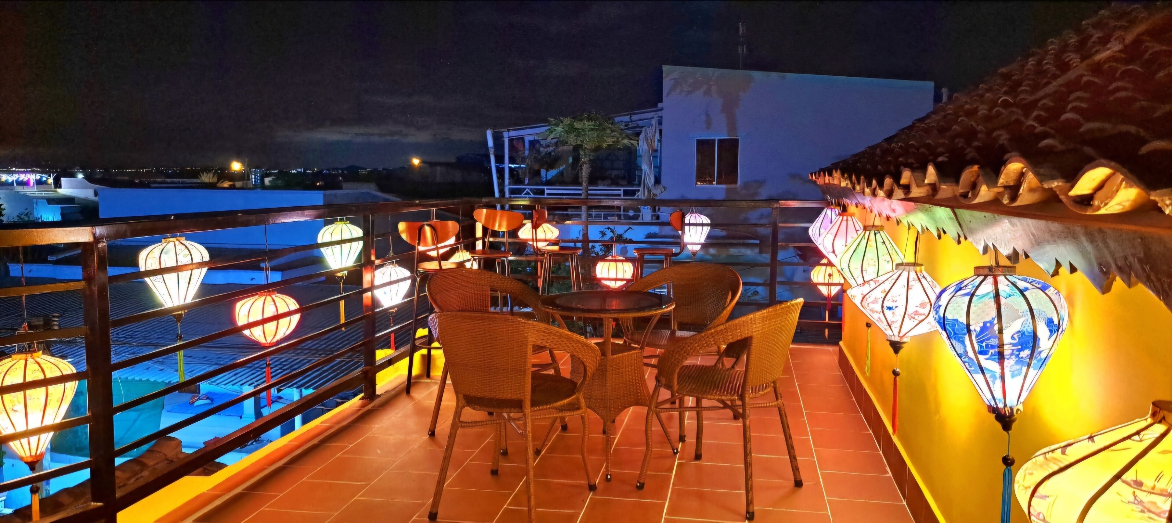 A rooftop terrace with wicker chairs and tables, illuminated by hanging lanterns.