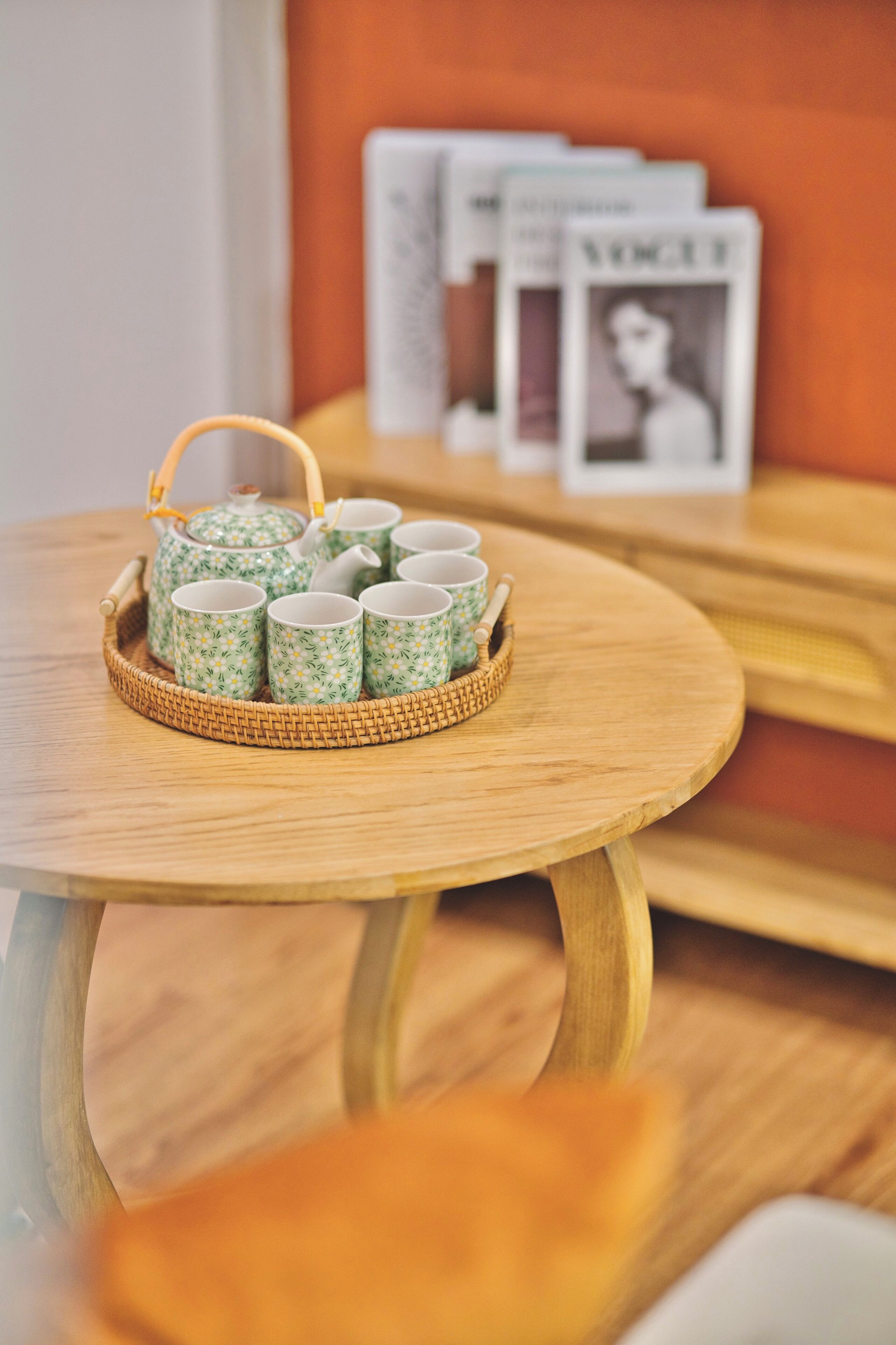 A round wooden table with a woven basket containing a teapot and six cups with floral patterns.