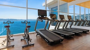 Et fitnesscenter med løbebånd, spinningcykler og romaskiner, med store vinduer og udsigt over havet.