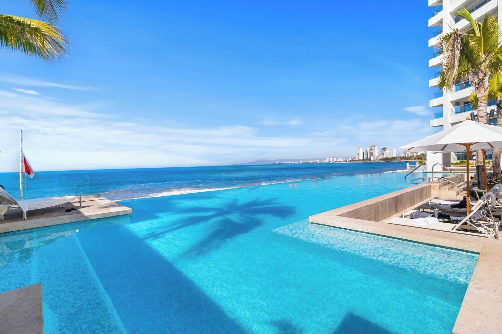 A clear blue pool with lounge chairs and umbrellas, overlooking a beach and city skyline.
