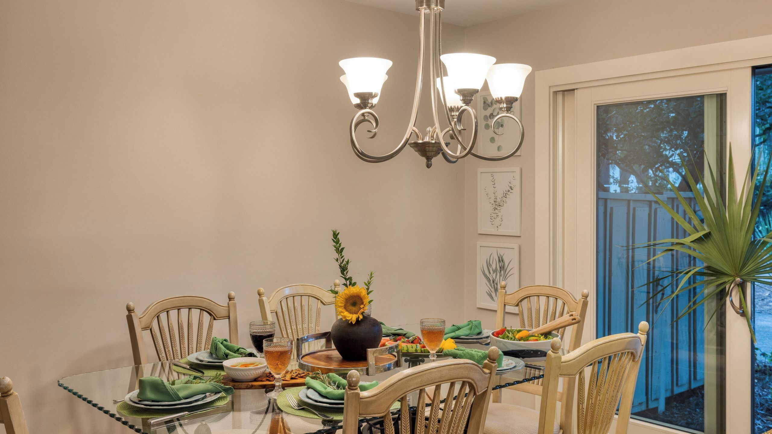 A dining area with a chandelier, wooden chairs, a glass table, and a window with a view.