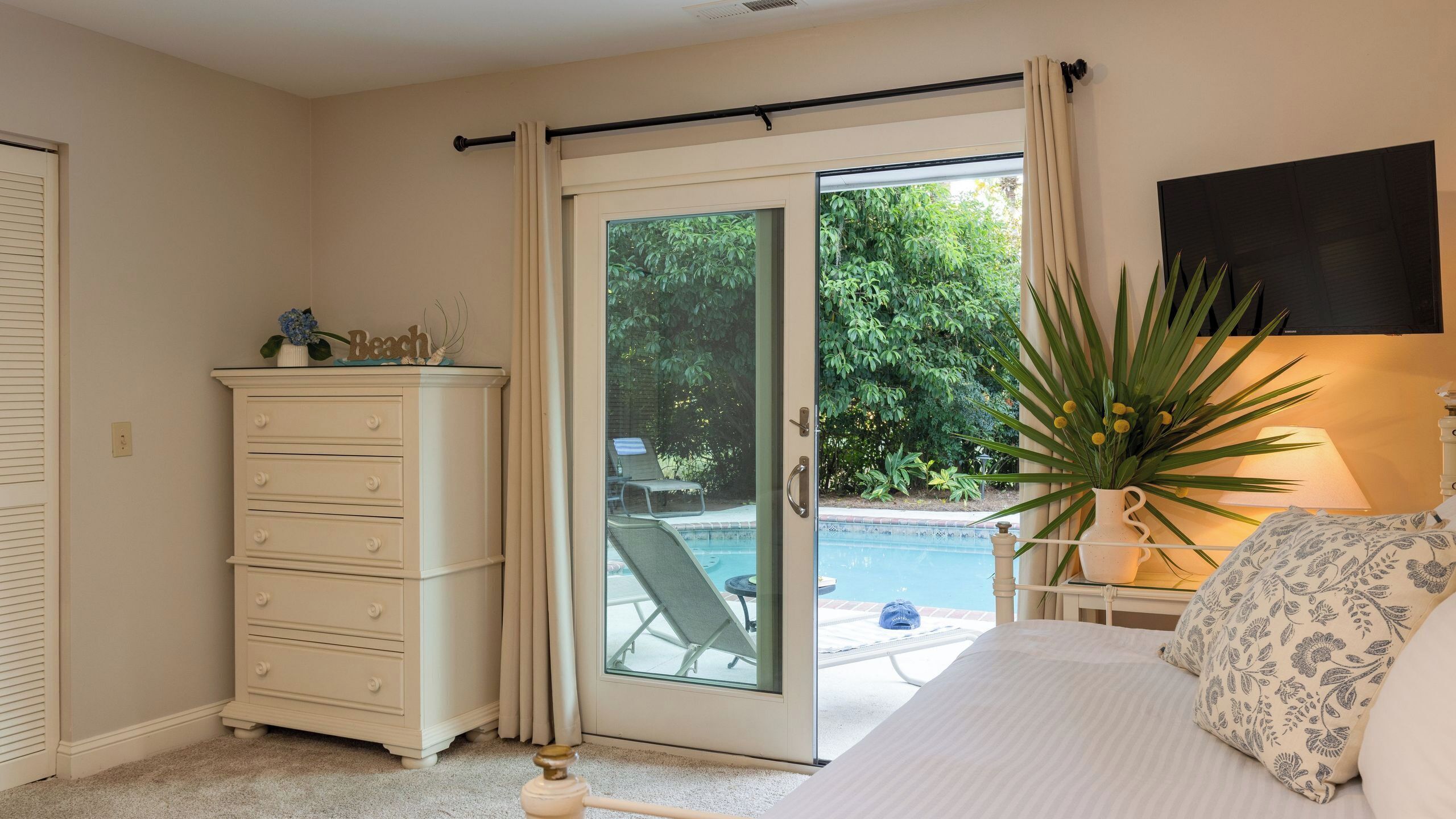 A bedroom with a bed, a dresser, a window with a view of a pool and greenery, and a lamp.