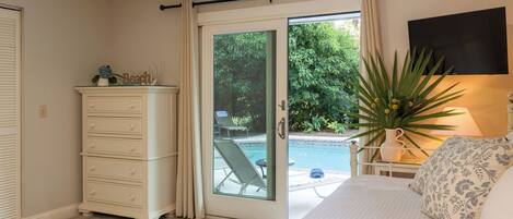A bedroom with a bed, a dresser, a window with a view of a pool and greenery, and a lamp.