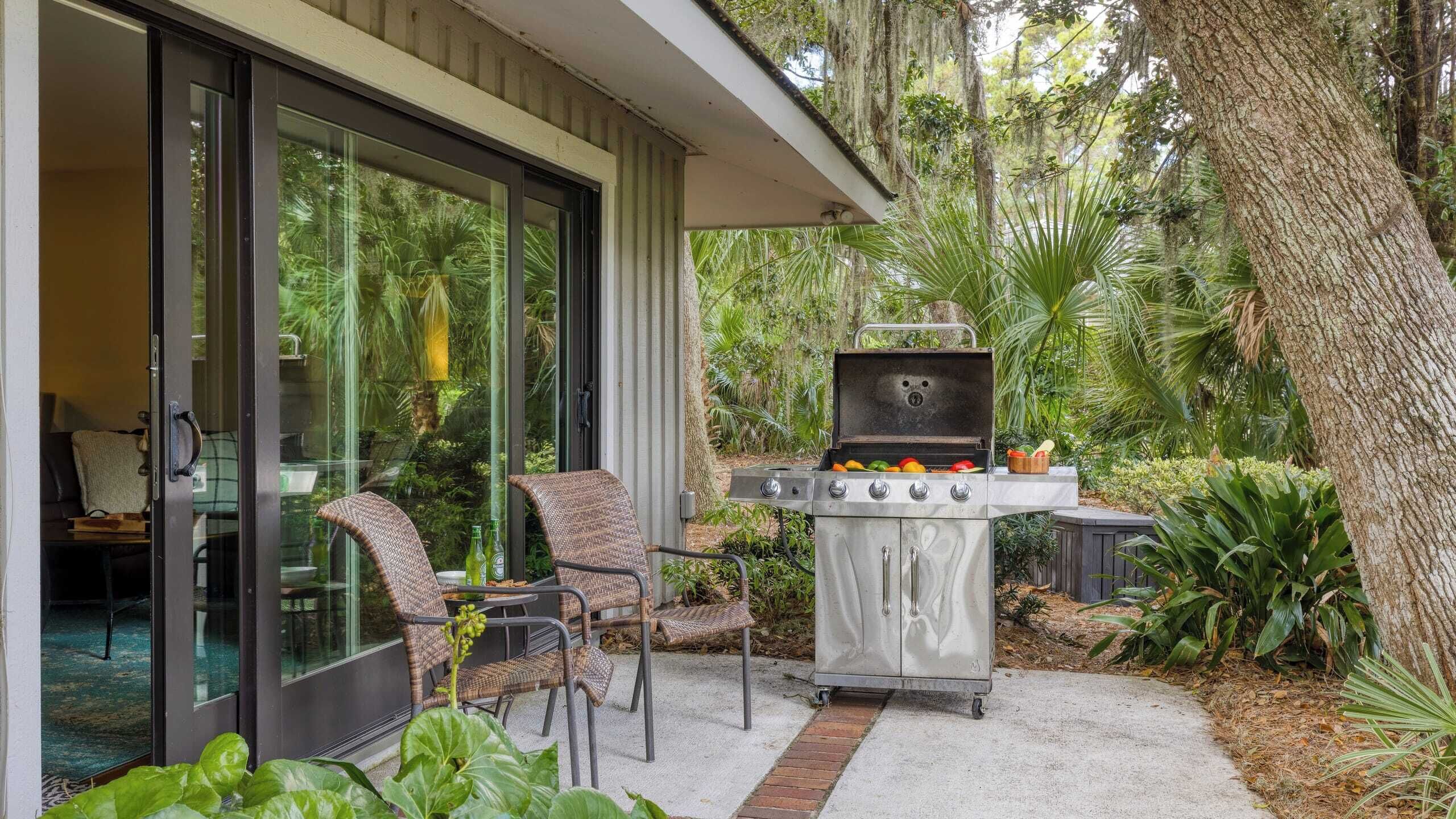 A patio with a grill, wicker chairs, and a glass door leading to a room.