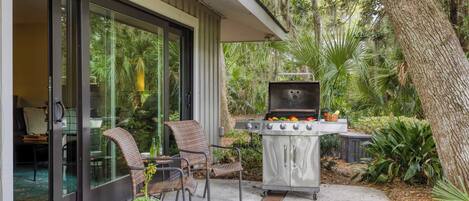 A patio with a grill, wicker chairs, and a glass door leading to a room.