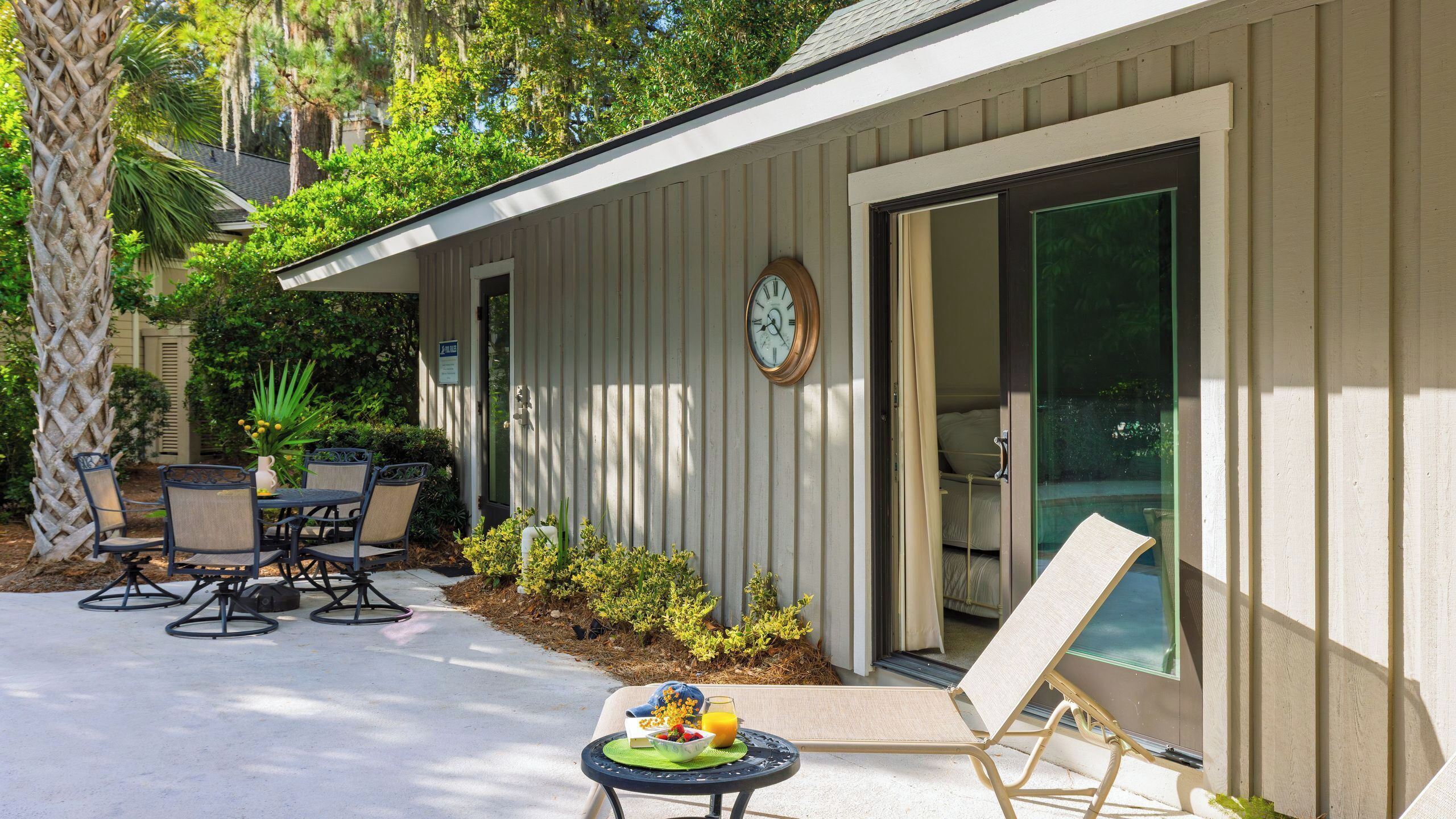 A house with a patio, outdoor seating, a clock, and a sliding glass door leading to a bedroom.