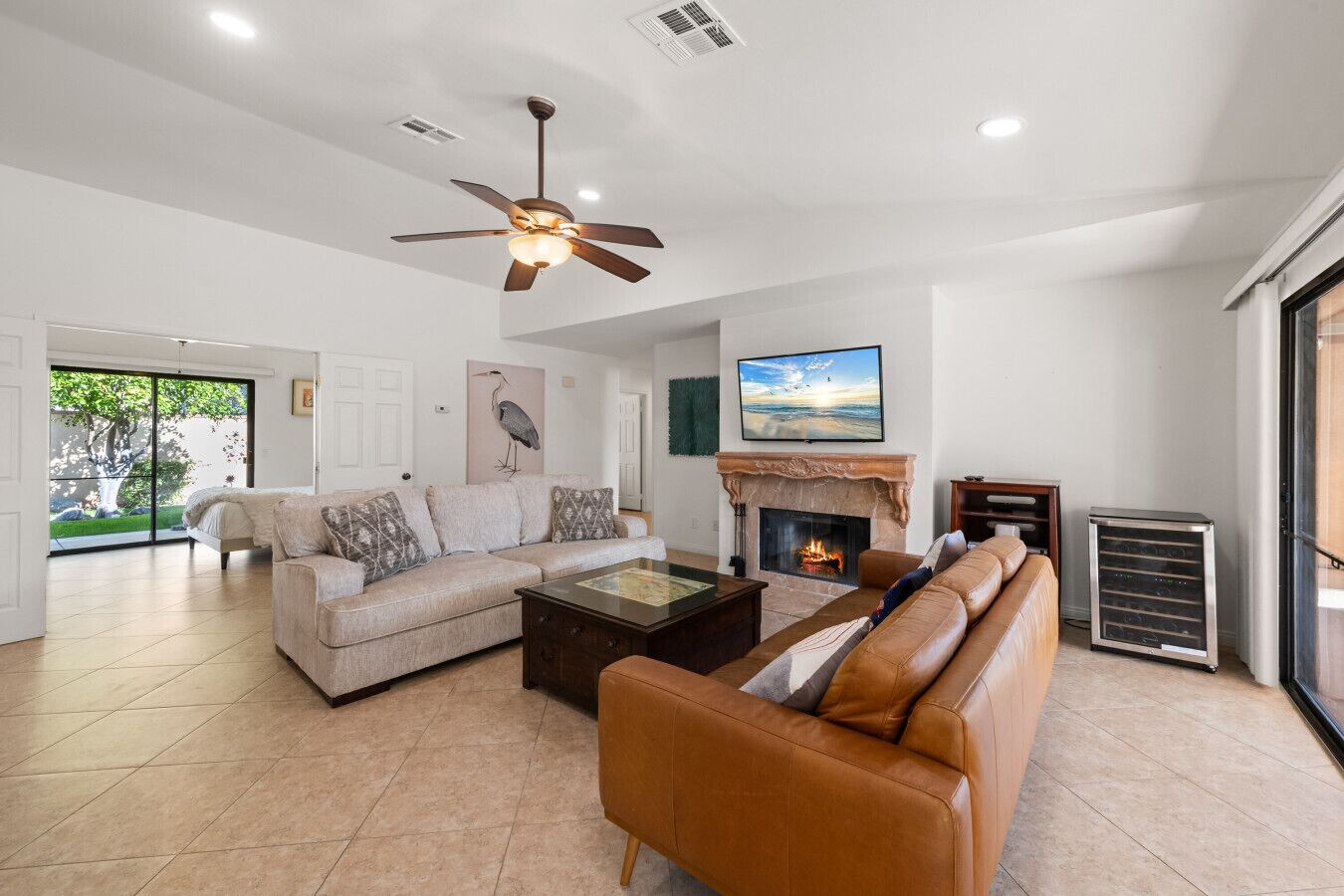 A spacious living room with a fireplace, a ceiling fan, and a large window overlooking a garden.