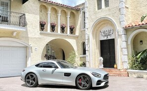 Une voiture de sport blanche est garée devant une maison de style méditerranéen, dotée d’un garage, de portes en arc de cercle et de colonnes décoratives.