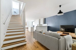 Uma sala de estar moderna com escada, um sofá cinza, uma televisão e uma cozinha ao fundo.