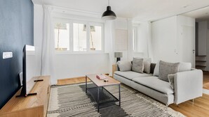Uma sala de estar moderna com um sofá cinzento, uma mesa de centro de madeira, um abajur preto e uma janela ampla com cortinas brancas.