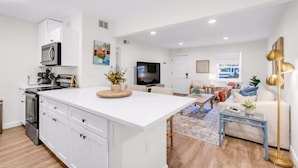 A modern kitchen with white cabinets, a central island, and a dining area with a blue coffee table. - Vibe at The Porter (Ocean Springs)