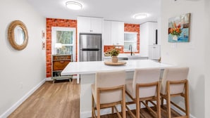 A modern kitchen with white cabinets, a stainless steel refrigerator, and a central island with chairs. - Vibe at The Porter (Ocean Springs)