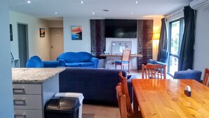 Uma sala de estar moderna com uma mesa de jantar de madeira, sofás azuis e uma TV fixada na parede.