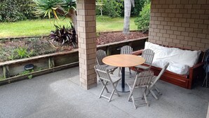 Um pátio externo com uma mesa redonda e quatro cadeiras, um banco de madeira com almofadas e uma parede de tijolos.