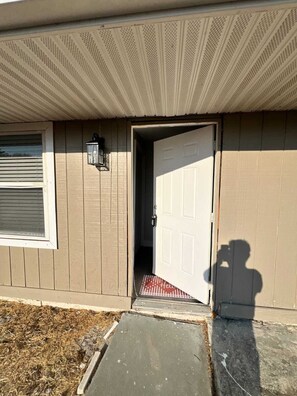 Une maison à l’extérieur beige, avec une porte blanche, une fenêtre équipée de stores et un petit porche surmonté d’un paillasson rouge.