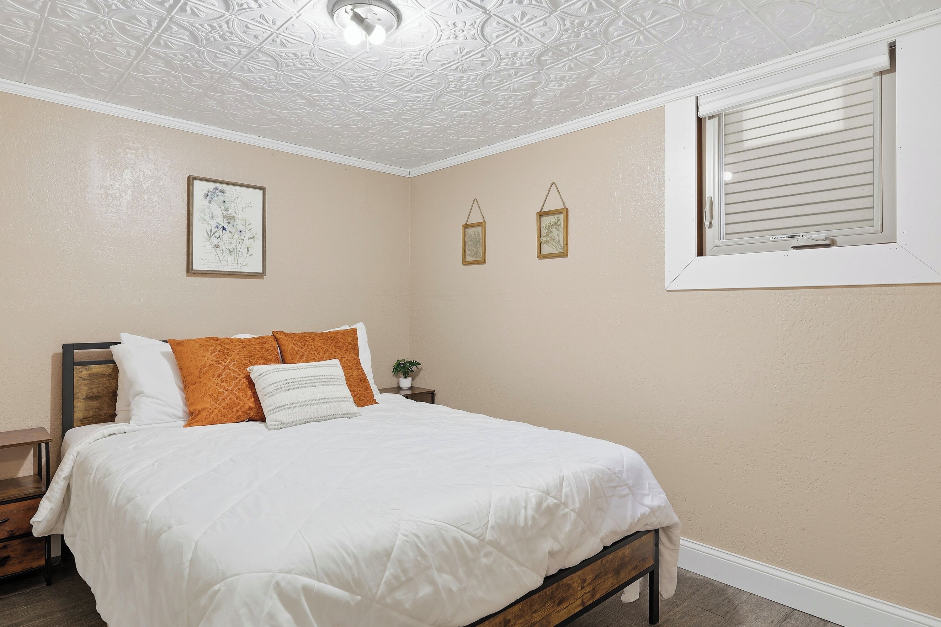 Une chambre à coucher comprenant un lit, une table de chevet, une fenêtre avec des stores et des œuvres d’art décoratives au mur.