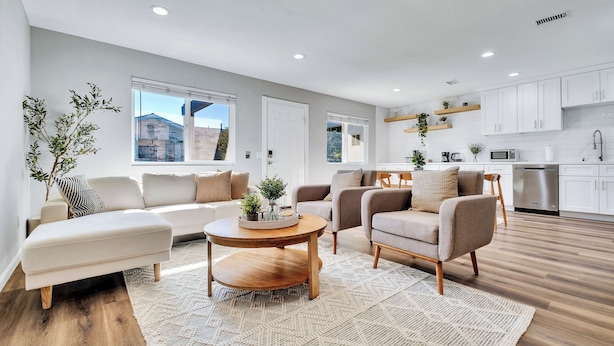 Una sala de estar moderna con un sofá, sillones, una mesa de centro y una cocina con armarios y electrodomésticos.