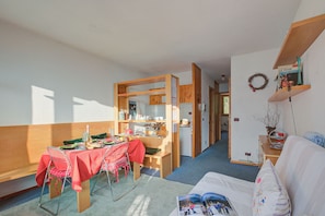 A compact room with a dining table, chairs, a sofa, a small kitchen area, and a bookshelf.
