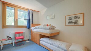 Une chambre avec un lit superposé, un lit simple, un bureau avec une chaise, un radiateur et une fenêtre avec des rideaux.