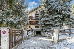 Un sentier enneigé mène à un immeuble à plusieurs étages doté de balcons, le tout étant entouré d’arbres chargés de neige.
