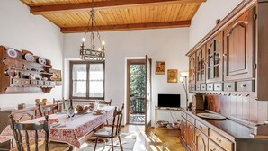 Een traditionele eetruimte met een houten tafel, stoelen en een kroonluchter. Er is een keuken met kasten en een televisie.