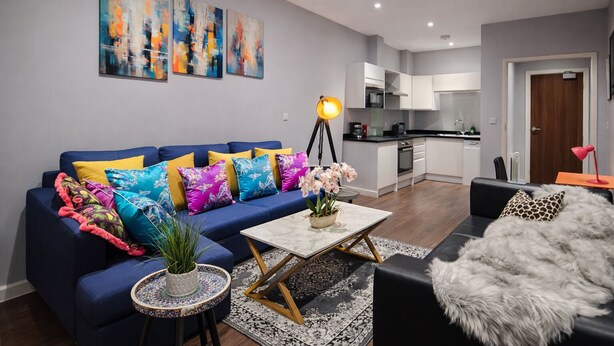 Ein modernes Wohnzimmer mit einem blauen Sofa, bunten Kissen, einem Couchtisch und einem kleinen Beistelltisch mit einer Pflanze. Im Hintergrund ist eine Küche mit weißen Schränken und einer schwarzen Arbeitsplatte zu sehen.