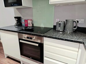 Een kitchenette met een kookplaat, oven en een magnetron. Op het aanrecht staan thee, suiker en koffie.