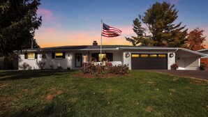 A single-story house with a front garage, a flagpole with an American flag, and a well-maintained lawn. - The Nook on the Farm—Pool • Hot Tub • Games (Pataskala)