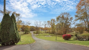 A paved driveway leading to a residential area with a well-maintained lawn, a red flowering bush, and a variety of trees. - The Nook on the Farm—Pool • Hot Tub • Games (Pataskala)