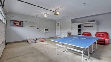 Um quarto com mesa de pingue-pongue, cadeiras de couro vermelho, alvo de dardos e um ventilador de teto.