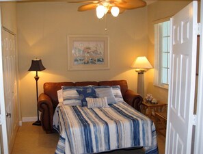 Un dormitorio con cama, dos lámparas, ventilador de techo, una obra de arte enmarcada y una puerta.