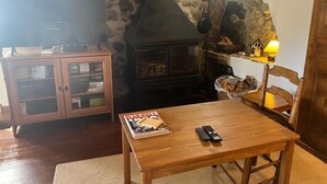 A cozy living room with a stone fireplace, a wooden table with a magazine and remotes, a television, and a bookshelf. - Country House with stunning views near Porte-Puymorens and Andorra (Carol)