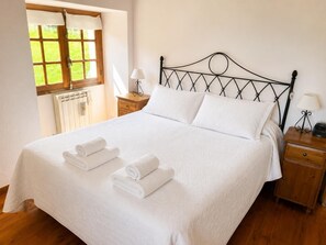 A neatly made bed with white linens and towels, a wooden nightstand with a lamp, and a window with a view of greenery.