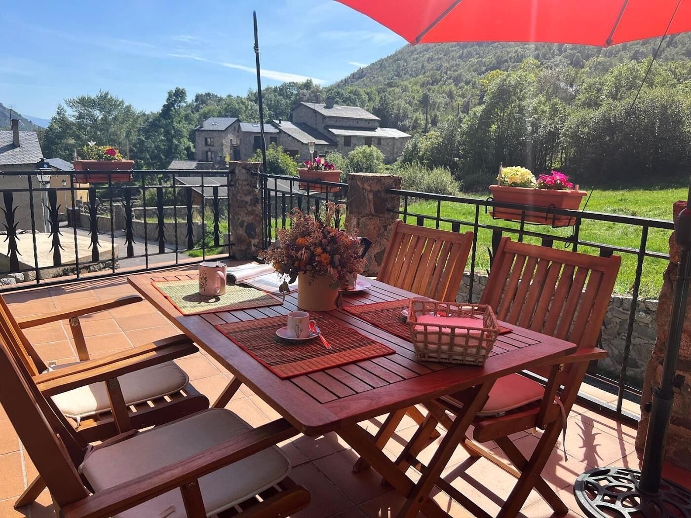 A wooden table and chairs on a balcony with a red umbrella, surrounded by potted plants and a view of houses and greenery.